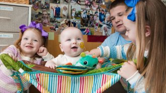 A smiling baby who got a personalized gene therapy is sitting in a multi-colored striped bouncy seat is surrounded by three siblings. A girl with long red hair and purple bows smiles at the camera on the left. A boy in a blue shirt watches as a girl with long blonde hair held back by a blue bow offers the baby a stuffed toy.
