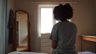 A woman with her back to the camera sits on a bed in a dimly lit room looking out the window