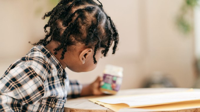 A young toddler wearing a flannel shirt sits at a table and holds a jar of food.