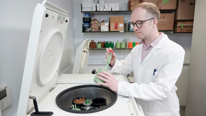 A scientist in a white lab coat operating a centrifuge machine in a laboratory, handling a green rotor with sample tubes, with lab equipment and storage boxes on shelves in the background.