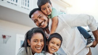 Smiling family of four outdoors, with a man giving a piggyback ride to a young boy, while a woman leans forward beside them, holding another smiling boy in front.