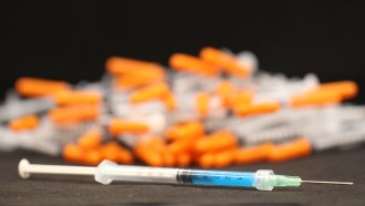 A syringe filled with blue hydrogel rests in front of a pile of orange-capped syringes.
