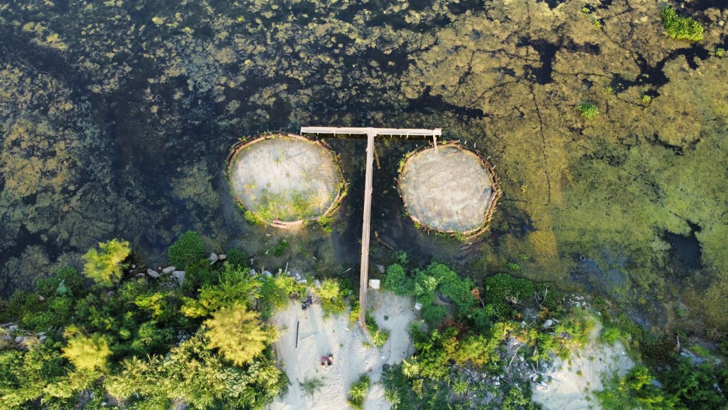 A birds-eye view of two small circular islands surrounded by marshy forest. The island on the right is more sparsely vegetated in this image.